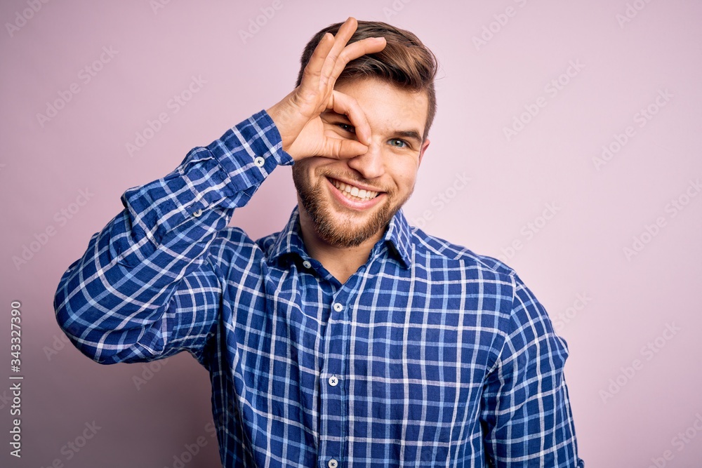Young handsome blond man with beard and blue eyes wearing casual shirt standing doing ok gesture with hand smiling, eye looking through fingers with happy face. Young handsome blond man with beard and blue eyes wearing casual shirt standing doing ok gesture with hand smiling, eye looking through fingers with happy face.