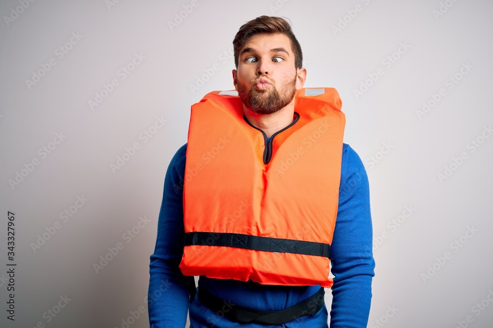 Fototapeta premium Young blond tourist man with beard and blue eyes wearing lifejacket over white background making fish face with lips, crazy and comical gesture. Funny expression.