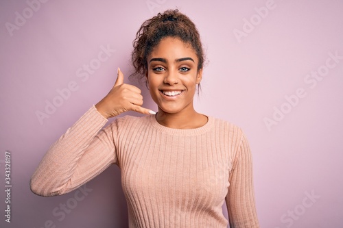 Young beautiful african american girl wearing casual sweater standing over pink background smiling doing phone gesture with hand and fingers like talking on the telephone. Communicating concepts.