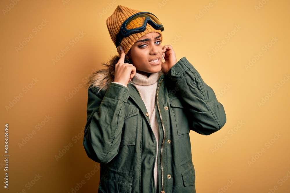 Young african american afro skier girl wearing snow sportswear and ski goggles covering ears with fingers with annoyed expression for the noise of loud music. Deaf concept. Young african american afro skier girl wearing snow sportswear and ski goggles covering ears with fingers with annoyed expression for the noise of loud music. Deaf concept.