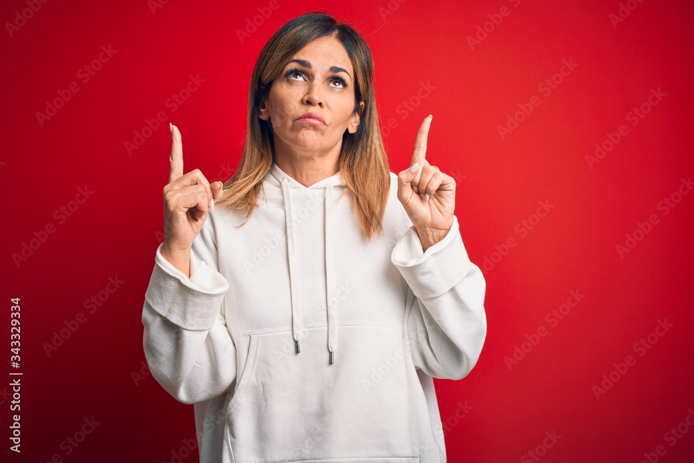 Middle age beautiful sportswoman wearing casual sweatshirt over isolated red background Pointing up looking sad and upset, indicating direction with fingers, unhappy and depressed. Middle age beautiful sportswoman wearing casual sweatshirt over isolated red background Pointing up looking sad and upset, indicating direction with fingers, unhappy and depressed.