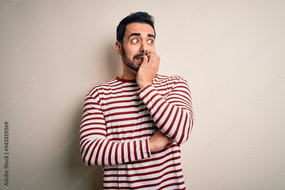 Young handsome man with beard wearing casual striped t-shirt standing over white background looking stressed and nervous with hands on mouth biting nails. Anxiety problem. Young handsome man with beard wearing casual striped t-shirt standing over white background looking stressed and nervous with hands on mouth biting nails. Anxiety problem.
