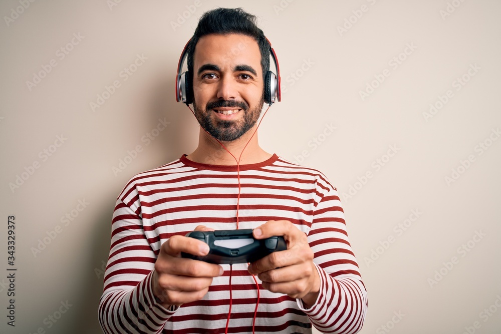 Young handsome gamer man with beard playing video game using joystick and headphones with a happy face standing and smiling with a confident smile showing teeth Young handsome gamer man with beard playing video game using joystick and headphones with a happy face standing and smiling with a confident smile showing teeth