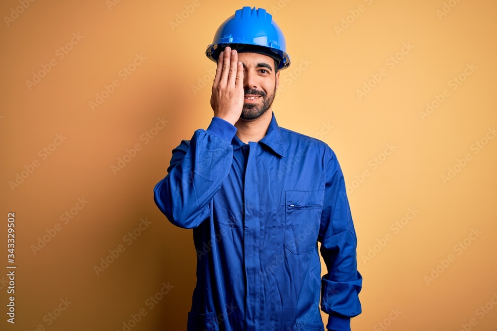 Mechanic man with beard wearing blue uniform and safety helmet over yellow background covering one eye with hand, confident smile on face and surprise emotion.
