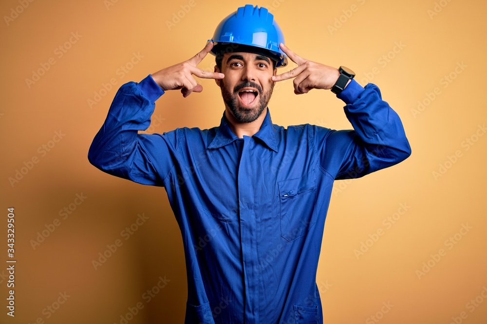 Mechanic man with beard wearing blue uniform and safety helmet over yellow background Doing peace symbol with fingers over face, smiling cheerful showing victory