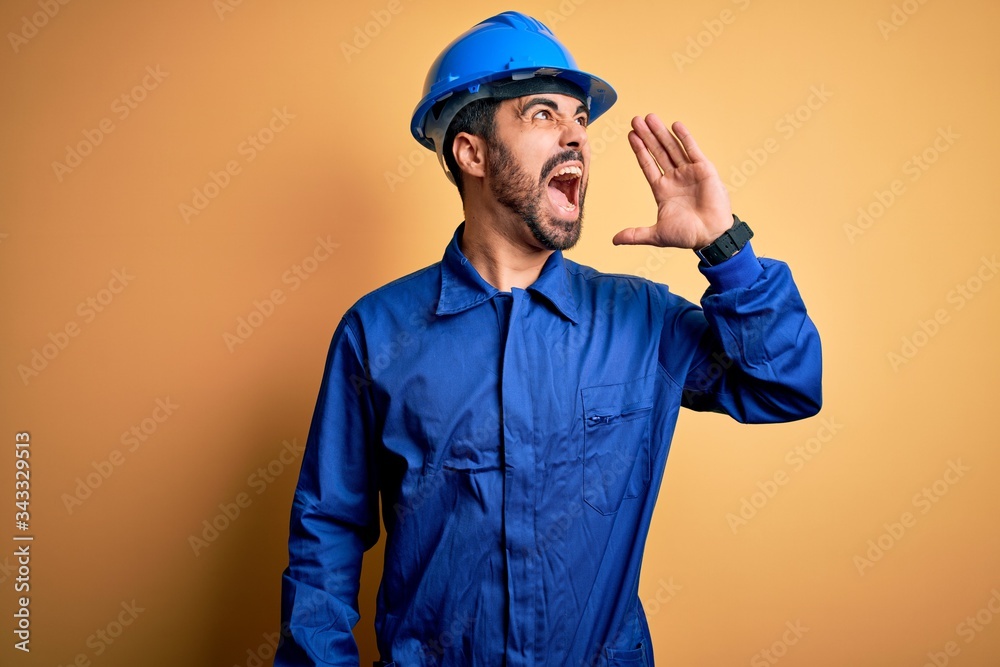 Mechanic man with beard wearing blue uniform and safety helmet over yellow background shouting and screaming loud to side with hand on mouth. Communication concept.