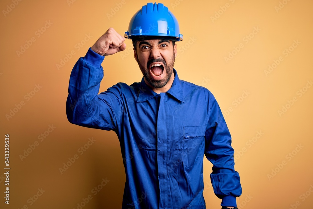 Mechanic man with beard wearing blue uniform and safety helmet over yellow background angry and mad raising fist frustrated and furious while shouting with anger. Rage and aggressive concept.