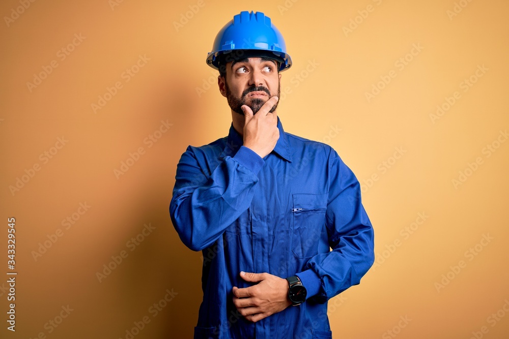 Mechanic man with beard wearing blue uniform and safety helmet over yellow background Thinking worried about a question, concerned and nervous with hand on chin