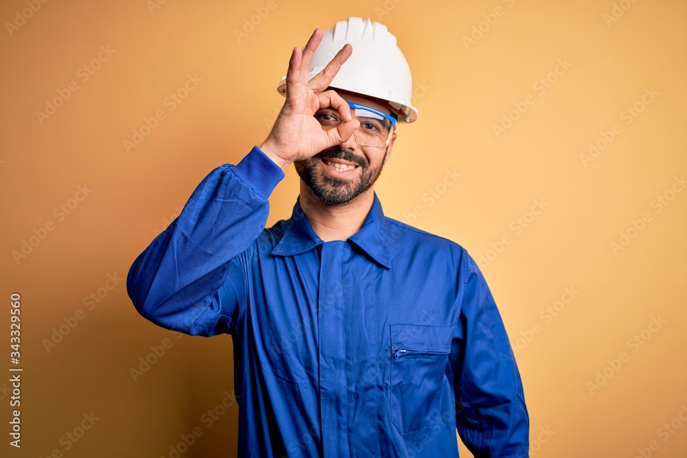 Mechanic man with beard wearing blue uniform and safety glasses over yellow background doing ok gesture with hand smiling, eye looking through fingers with happy face. Mechanic man with beard wearing blue uniform and safety glasses over yellow background doing ok gesture with hand smiling, eye looking through fingers with happy face.