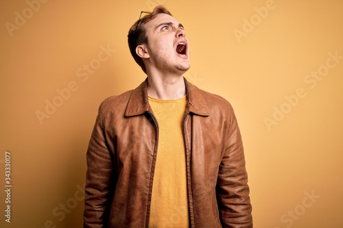 Young handsome redhead man wearing casual leather jacket over isolated yellow background angry and mad screaming frustrated and furious, shouting with anger. Rage and aggressive concept.