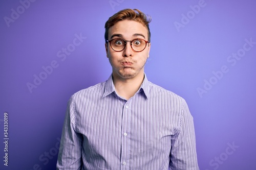 Young handsome redhead man wearing casual shirt and glasses over purple background puffing cheeks with funny face. Mouth inflated with air, crazy expression.