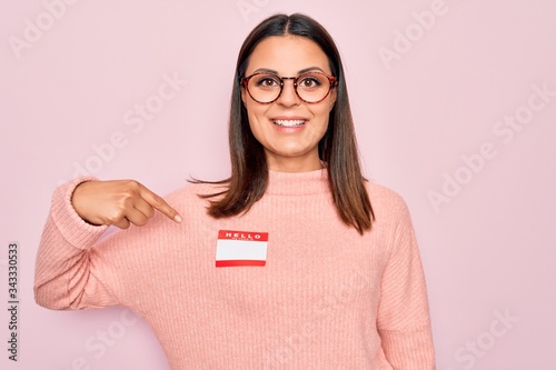 Young beautiful brunette woman wearing sticker with hello my name is message Smiling happy pointing with hand and finger