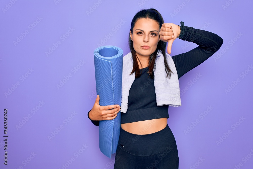 Young beautiful brunette sporty woman using sport towel hoding mat to do yoga with angry face, negative sign showing dislike with thumbs down, rejection concept Young beautiful brunette sporty woman using sport towel hoding mat to do yoga with angry face, negative sign showing dislike with thumbs down, rejection concept