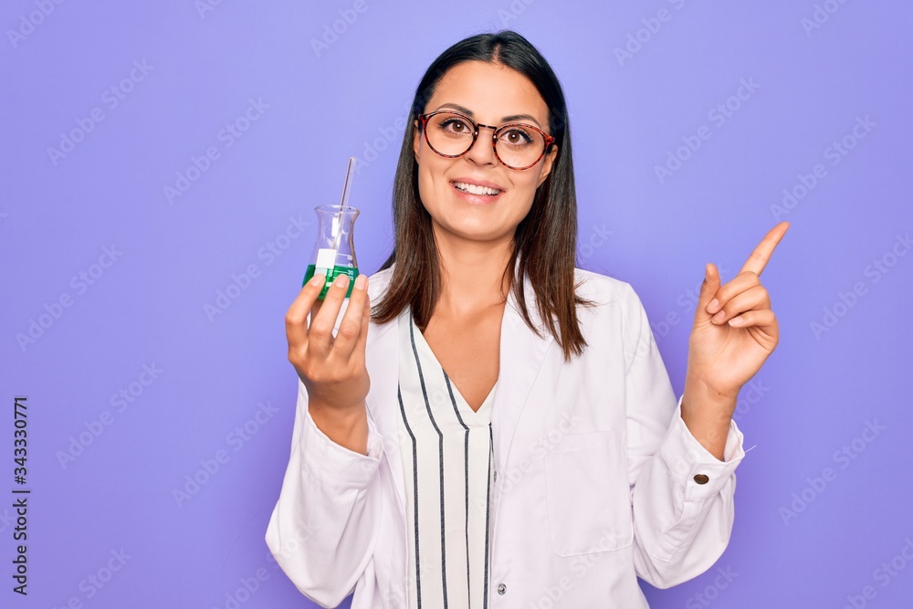 Young beautiful brunette scientist woman wearing coat and glasses holding test tube Smiling happy pointing with hand and finger to the side Young beautiful brunette scientist woman wearing coat and glasses holding test tube Smiling happy pointing with hand and finger to the side