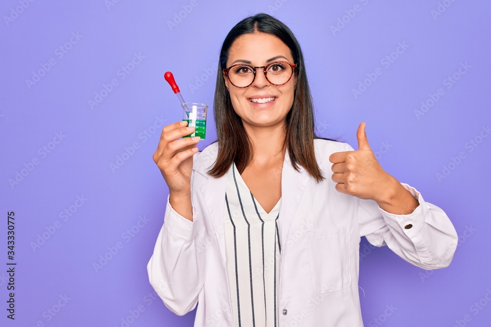 Young beautiful brunette scientist woman wearing coat and glasses holding test tube Smiling happy and positive, thumb up doing excellent and approval sign Young beautiful brunette scientist woman wearing coat and glasses holding test tube Smiling happy and positive, thumb up doing excellent and approval sign