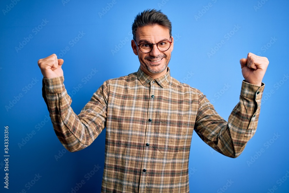 Naklejka premium Young handsome man wearing casual shirt and glasses standing over blue background celebrating surprised and amazed for success with arms raised and open eyes. Winner concept.