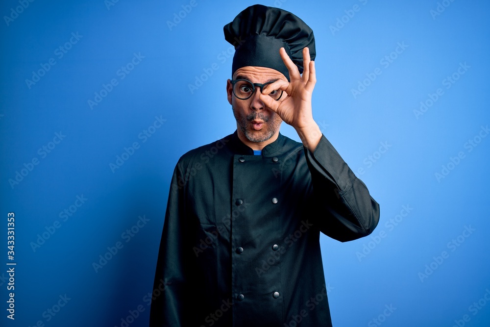 Young handsome chef man wearing cooker uniform and hat over isolated blue background doing ok gesture shocked with surprised face, eye looking through fingers. Unbelieving expression. Young handsome chef man wearing cooker uniform and hat over isolated blue background doing ok gesture shocked with surprised face, eye looking through fingers. Unbelieving expression.