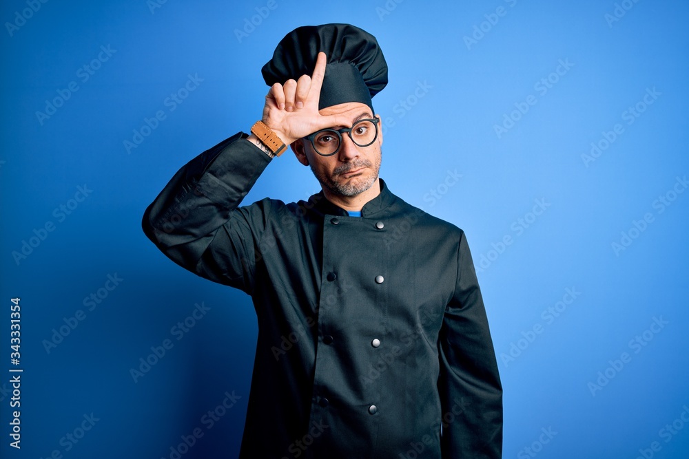 Young handsome chef man wearing cooker uniform and hat over isolated blue background making fun of people with fingers on forehead doing loser gesture mocking and insulting.