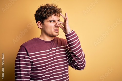 Young blond handsome man with curly hair wearing casual striped sweater smelling something stinky and disgusting  intolerable smell  holding breath with fingers on nose. Bad smell