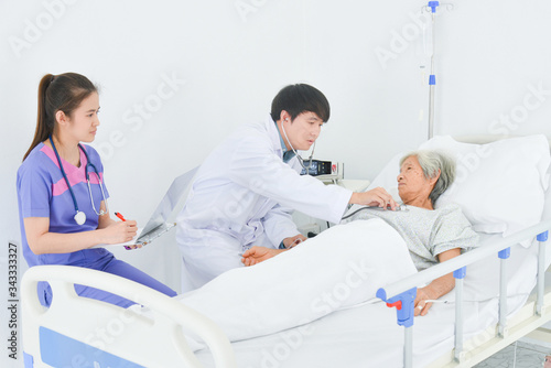 Doctor using stethoscope to listen heartbeat of senior asian female, patient lying on bed in hospital ward.
