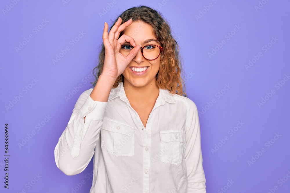 Young beautiful woman with blue eyes wearing casual shirt and glasses over purple background doing ok gesture with hand smiling, eye looking through fingers with happy face.