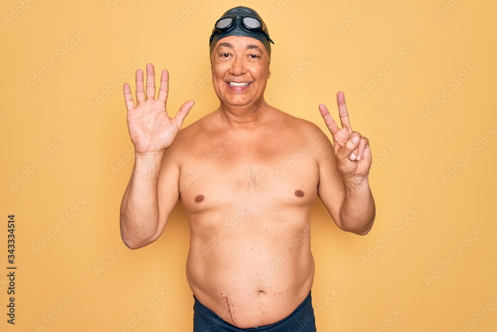 Middle age senior grey-haired swimmer man wearing swimsuit, cap and goggles showing and pointing up with fingers number seven while smiling confident and happy.