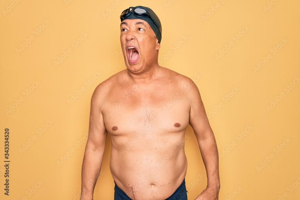 Middle age senior grey-haired swimmer man wearing swimsuit, cap and goggles angry and mad screaming frustrated and furious, shouting with anger. Rage and aggressive concept.