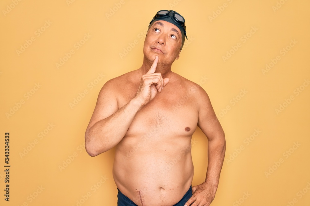 Middle age senior grey-haired swimmer man wearing swimsuit, cap and goggles Thinking concentrated about doubt with finger on chin and looking up wondering