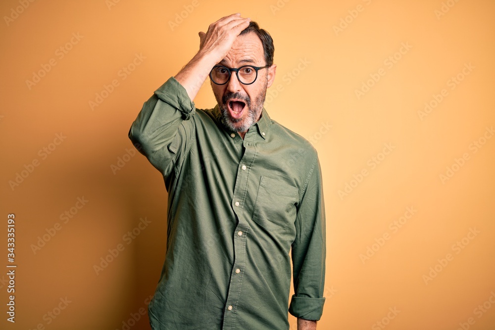 Middle age hoary man wearing casual green shirt and glasses over isolated yellow background surprised with hand on head for mistake, remember error. Forgot, bad memory concept. Middle age hoary man wearing casual green shirt and glasses over isolated yellow background surprised with hand on head for mistake, remember error. Forgot, bad memory concept.