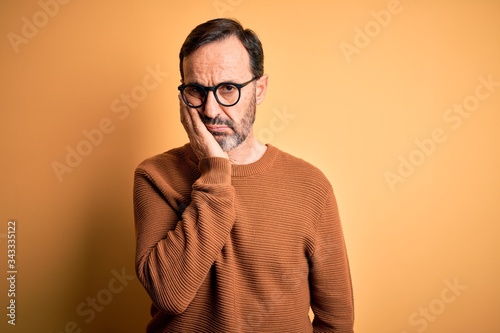 Middle age hoary man wearing brown sweater and glasses over isolated yellow background thinking looking tired and bored with depression problems with crossed arms.