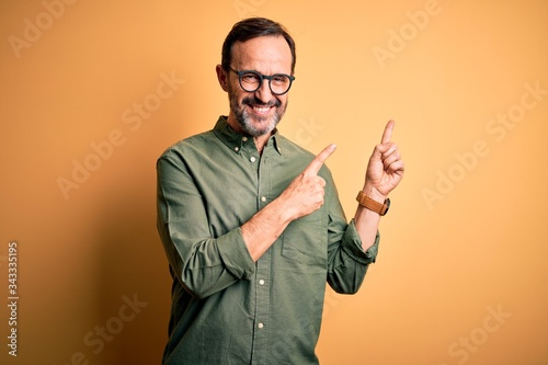 Canvas Print Middle age hoary man wearing casual green shirt and glasses over isolated yellow background smiling and looking at the camera pointing with two hands and fingers to the side