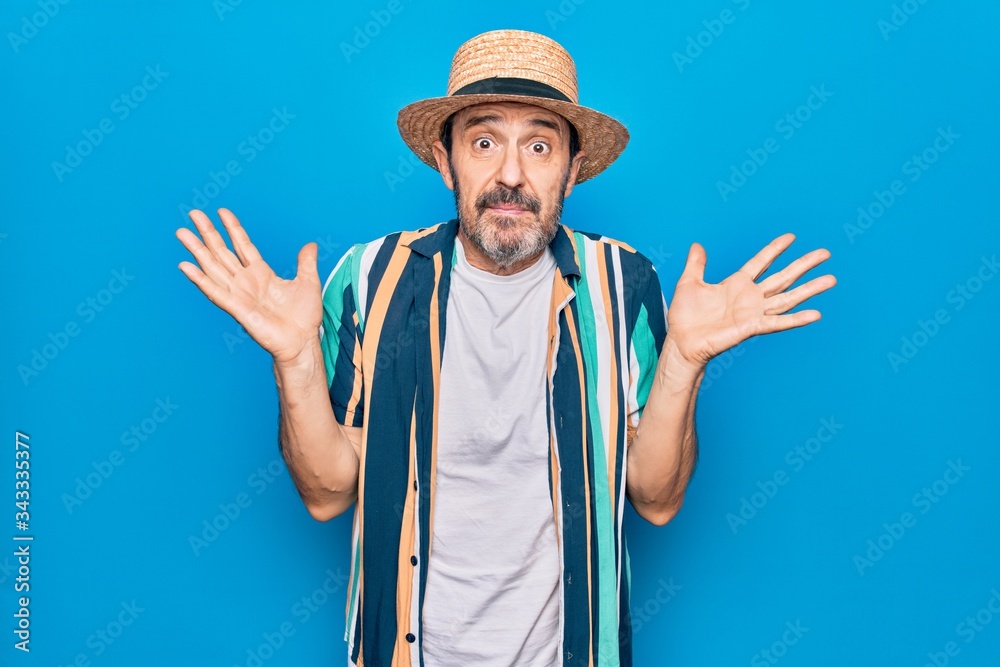 Middle age handsome tourist man on vacation wearing shirt and hat over blue background clueless and confused with open arms, no idea and doubtful face.
