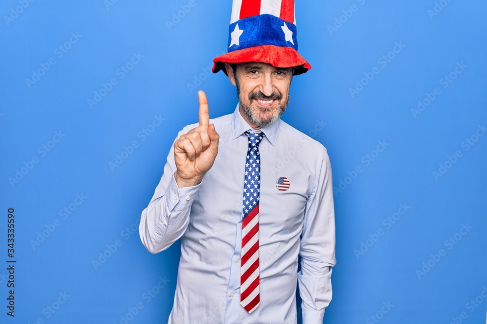 Middle age handsome patriotic man wearing united states hat and tie over blue background smiling with an idea or question pointing finger up with happy face, number one Middle age handsome patriotic man wearing united states hat and tie over blue background smiling with an idea or question pointing finger up with happy face, number one