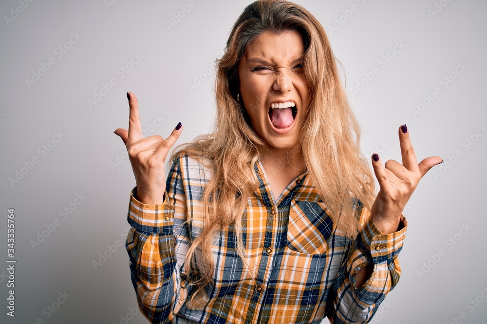 Young beautiful blonde woman wearing casual shirt standing over isolated white background shouting with crazy expression doing rock symbol with hands up. Music star. Heavy concept. Young beautiful blonde woman wearing casual shirt standing over isolated white background shouting with crazy expression doing rock symbol with hands up. Music star. Heavy concept.