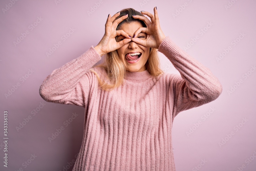 Young beautiful blonde woman wearing casual pink sweater over isolated background doing ok gesture like binoculars sticking tongue out, eyes looking through fingers. Crazy expression. Young beautiful blonde woman wearing casual pink sweater over isolated background doing ok gesture like binoculars sticking tongue out, eyes looking through fingers. Crazy expression.