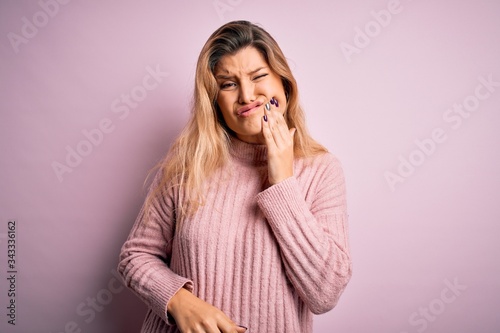 Young beautiful blonde woman wearing casual pink sweater over isolated background touching mouth with hand with painful expression because of toothache or dental illness on teeth. Dentist © Krakenimages.com