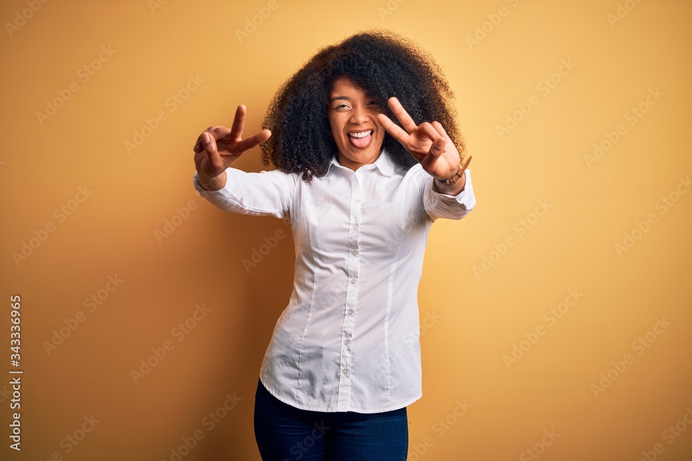 Young beautiful african american elegant woman with afro hair standing over yellow background smiling with tongue out showing fingers of both hands doing victory sign. Number two. Young beautiful african american elegant woman with afro hair standing over yellow background smiling with tongue out showing fingers of both hands doing victory sign. Number two.