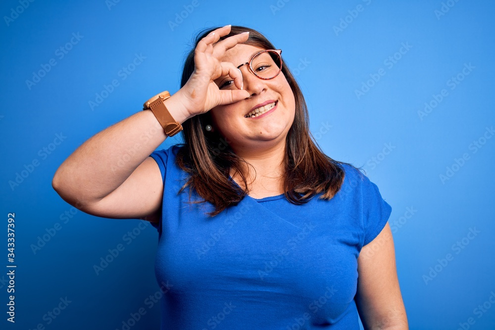 Beautiful brunette plus size woman wearing casual t-shirt over isolated blue background doing ok gesture with hand smiling, eye looking through fingers with happy face.