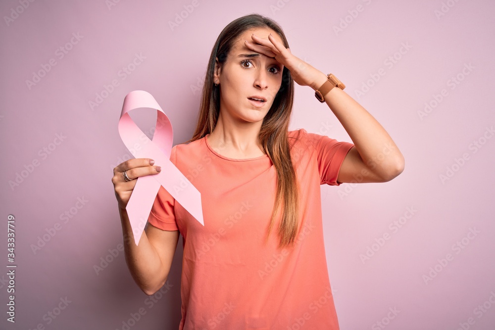 Young beautiful woman holding pink cancer ribbon standing over isolated pink background stressed with hand on head, shocked with shame and surprise face, angry and frustrated. Fear and upset Young beautiful woman holding pink cancer ribbon standing over isolated pink background stressed with hand on head, shocked with shame and surprise face, angry and frustrated. Fear and upset