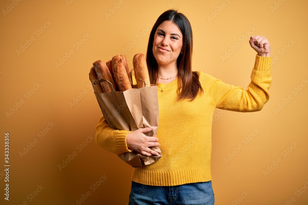 Young beautiful woman holding a bag of fresh healthy bread over yellow background Strong person showing arm muscle, confident and proud of power Young beautiful woman holding a bag of fresh healthy bread over yellow background Strong person showing arm muscle, confident and proud of power
