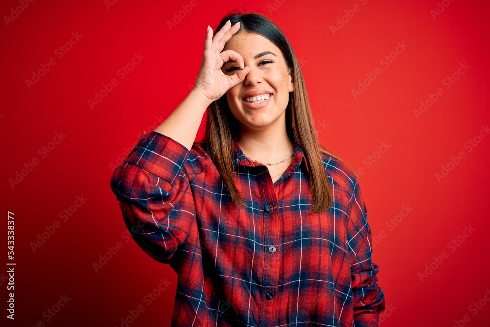 Young beautiful woman wearing casual shirt over red background doing ok gesture with hand smiling, eye looking through fingers with happy face. Young beautiful woman wearing casual shirt over red background doing ok gesture with hand smiling, eye looking through fingers with happy face.