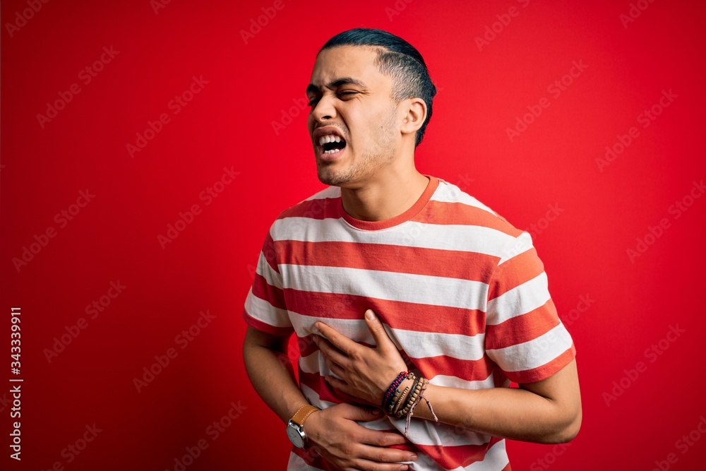 Young brazilian man wearing casual striped t-shirt standing over isolated red background with hand on stomach because nausea, painful disease feeling unwell. Ache concept. Young brazilian man wearing casual striped t-shirt standing over isolated red background with hand on stomach because nausea, painful disease feeling unwell. Ache concept.