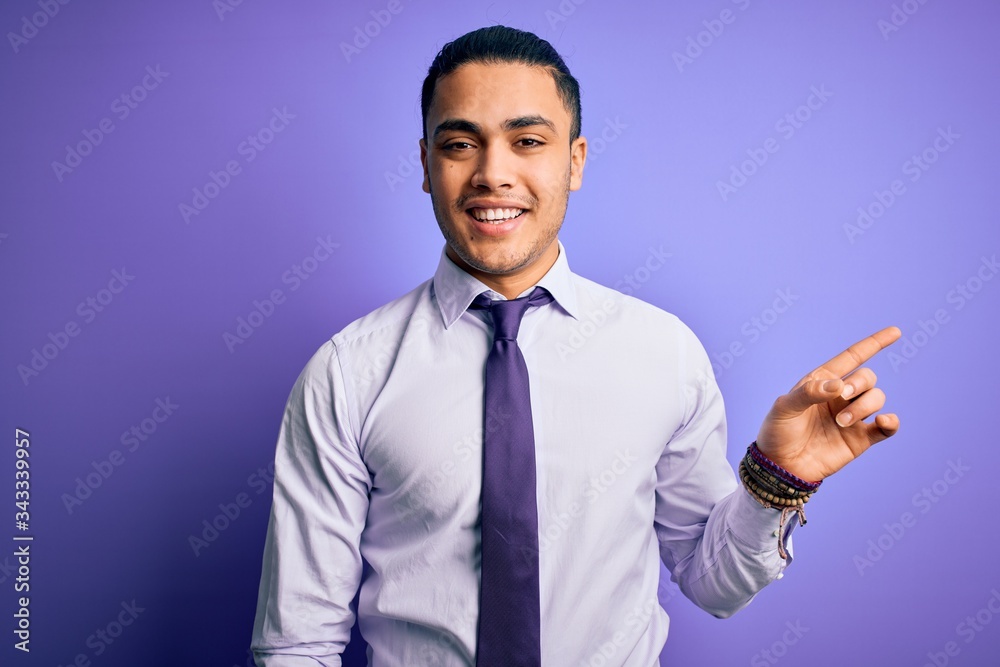 Young brazilian businessman wearing elegant tie standing over isolated purple background with a big smile on face, pointing with hand finger to the side looking at the camera. Young brazilian businessman wearing elegant tie standing over isolated purple background with a big smile on face, pointing with hand finger to the side looking at the camera.
