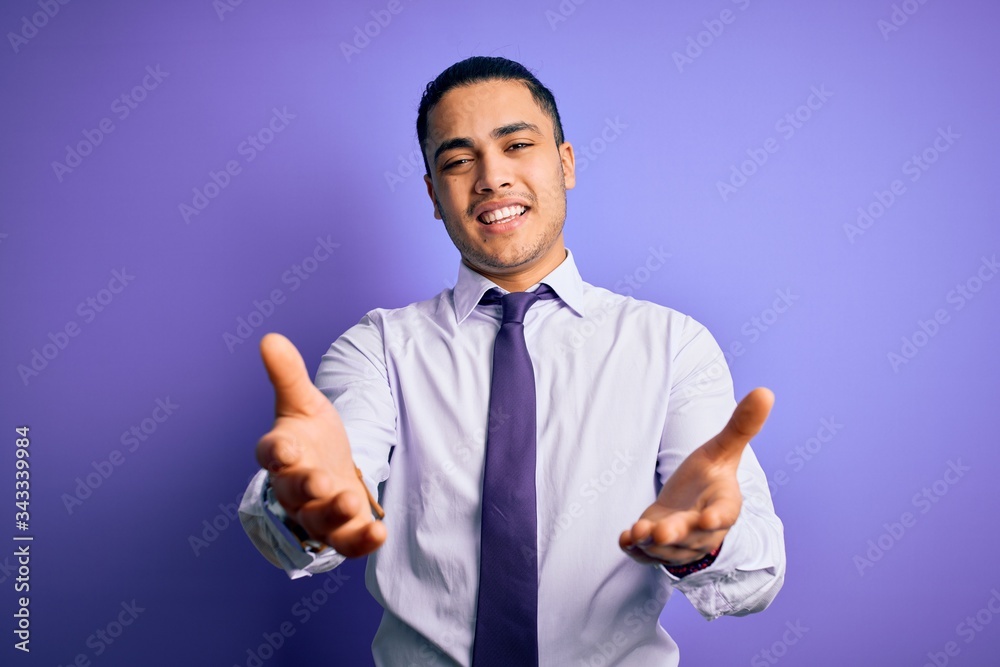 Young brazilian businessman wearing elegant tie standing over isolated purple background smiling cheerful offering hands giving assistance and acceptance. Young brazilian businessman wearing elegant tie standing over isolated purple background smiling cheerful offering hands giving assistance and acceptance.