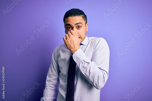 Young brazilian businessman wearing elegant tie standing over isolated purple background smelling something stinky and disgusting, intolerable smell, holding breath with fingers on nose. Bad smell © Krakenimages.com