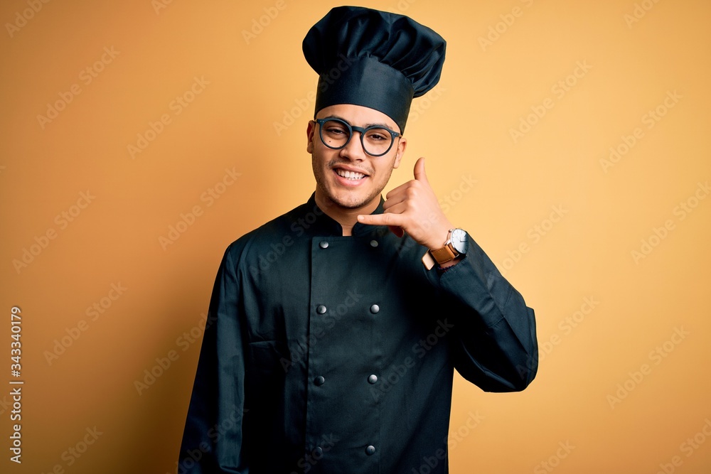Young brazilian chef man wearing cooker uniform and hat over isolated yellow background smiling doing phone gesture with hand and fingers like talking on the telephone. Communicating concepts. Young brazilian chef man wearing cooker uniform and hat over isolated yellow background smiling doing phone gesture with hand and fingers like talking on the telephone. Communicating concepts.