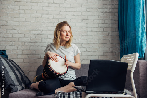 Life in coronavirus quarantine, education and activities at home during lockdown. Pretty young woman makes music playing the Djembe, looking at laptop. Remote online course of playing the drum.