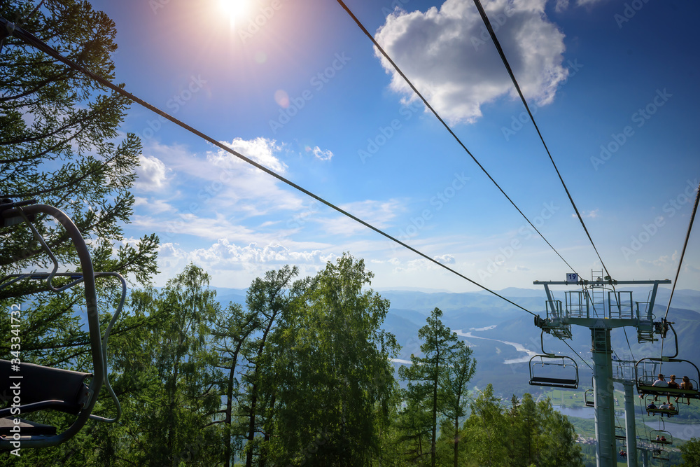 Ski lift in the mountains on sunny day against blue sky, white clouds, green hills and mountain lake. Mountain valley with cable car, view from top. Ski resort in summer.