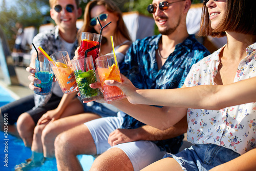 Close view. Friends clinking glasses with fresh colorful cocktails sitting by swimming pool on sunny summer day. People toast drinking beverages at luxury villa poolside party on tropical vacation.