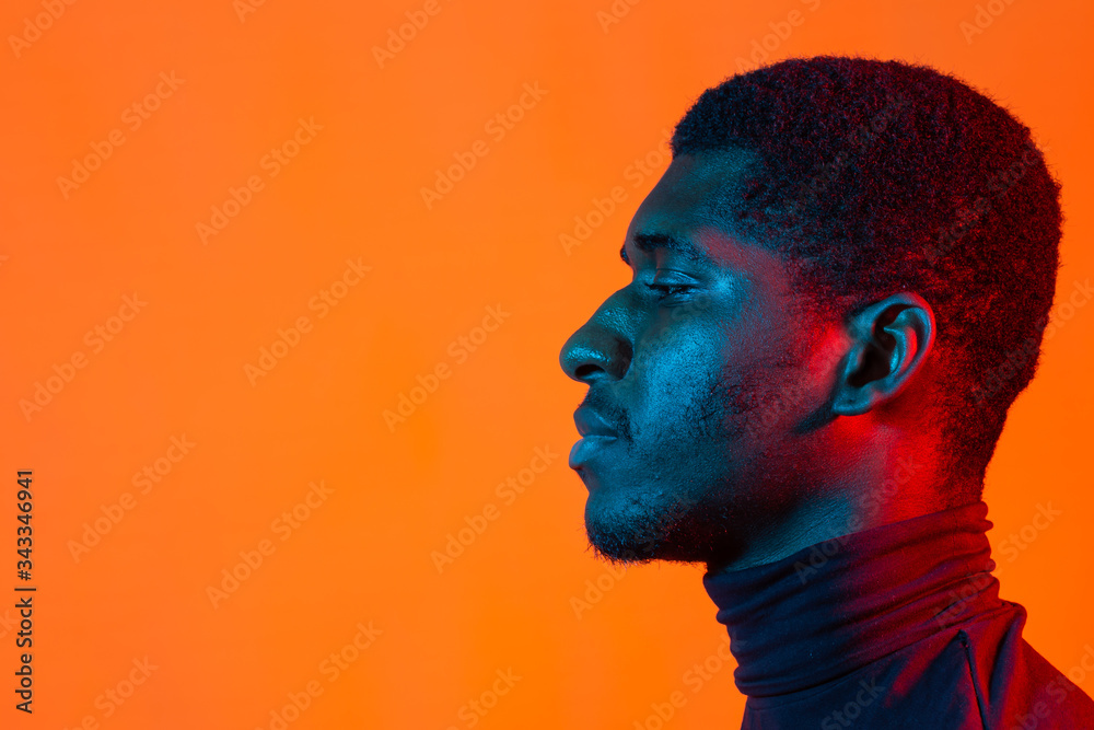 Young african-american man in neon light. Male portrait side view. Concept of human emotions, facial expression. Orange background with copy space.